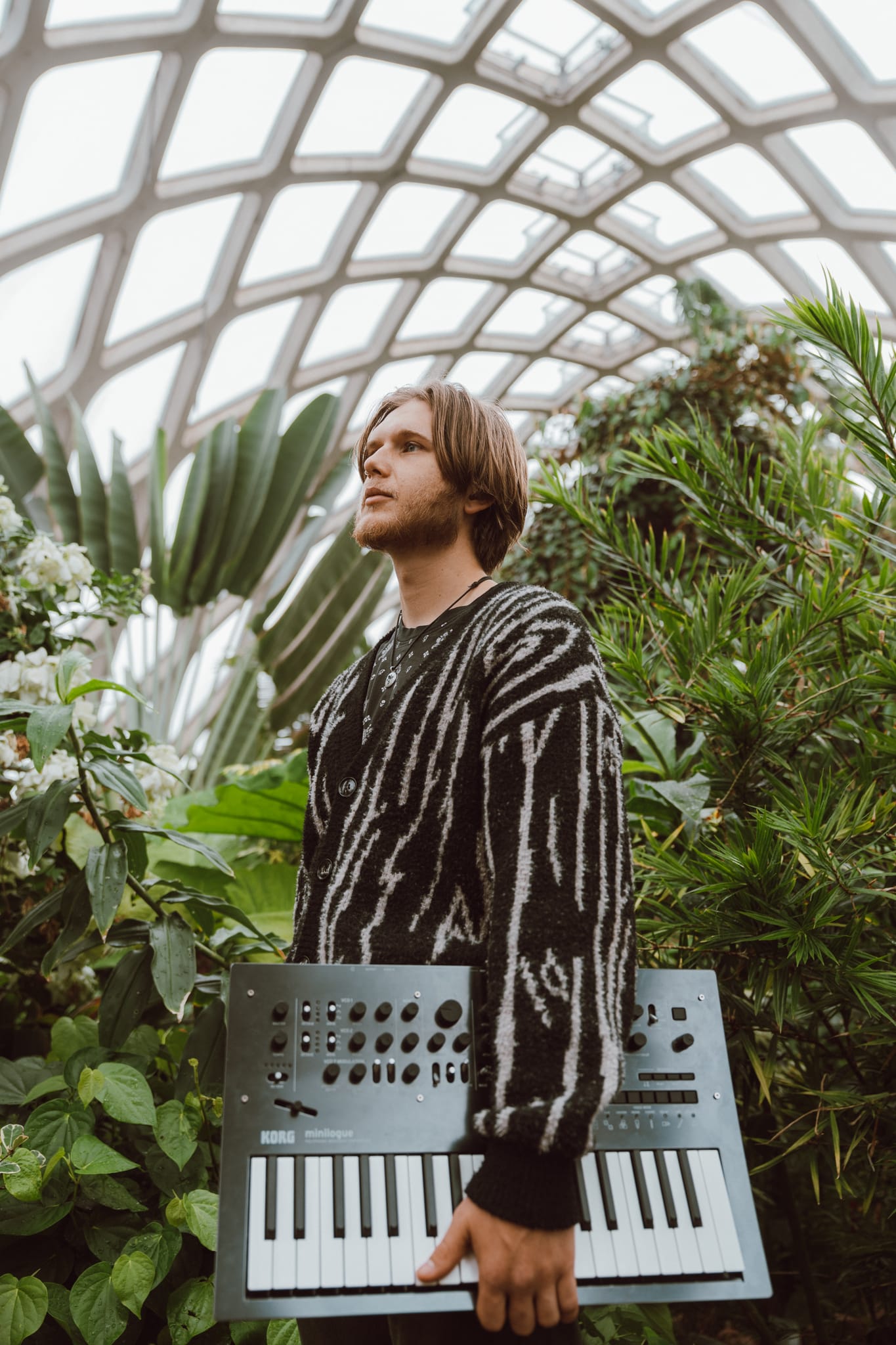Abe standing in a glass-domed conservatory surrounded by tropical plants, holding a synthesizer