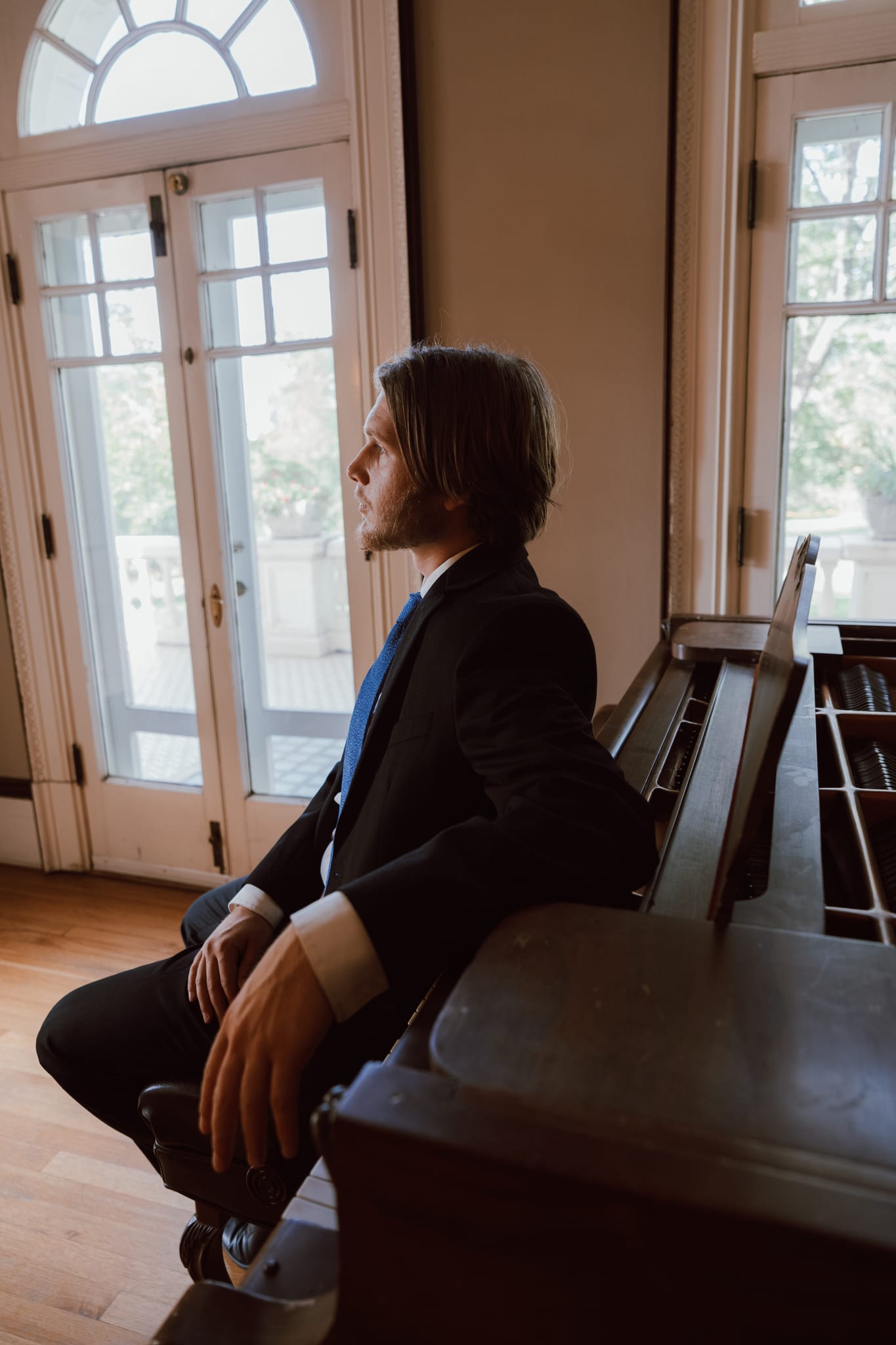 Abe in a dark suit leaning back against an upright piano in a sunlit room with arched windows