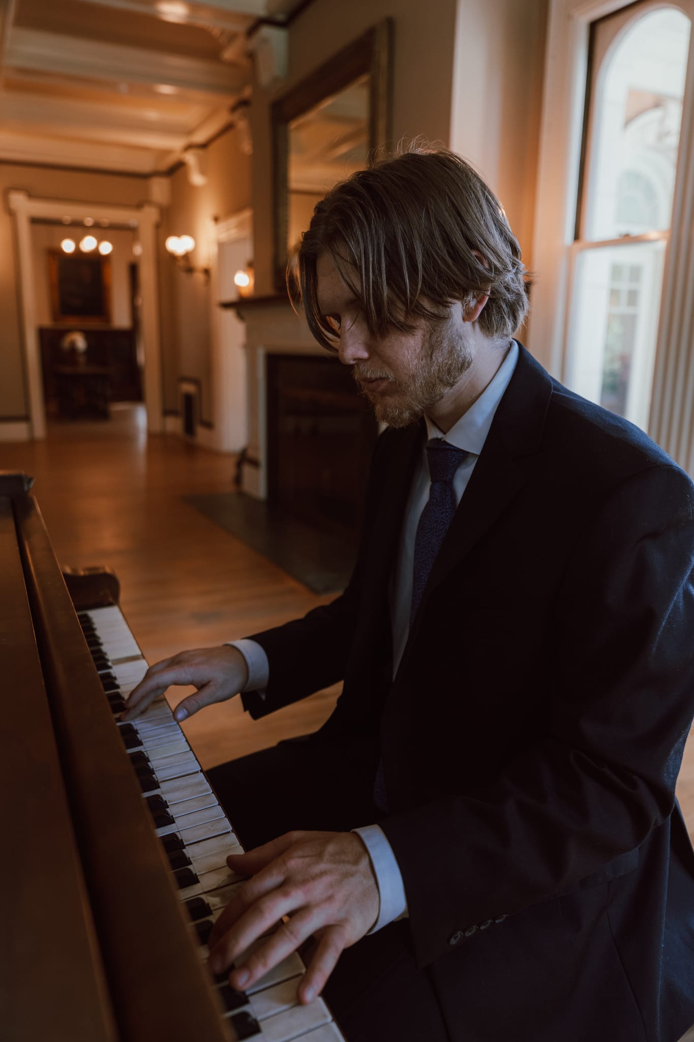 Abe in a dark suit playing an upright piano in a wood-floored hallway