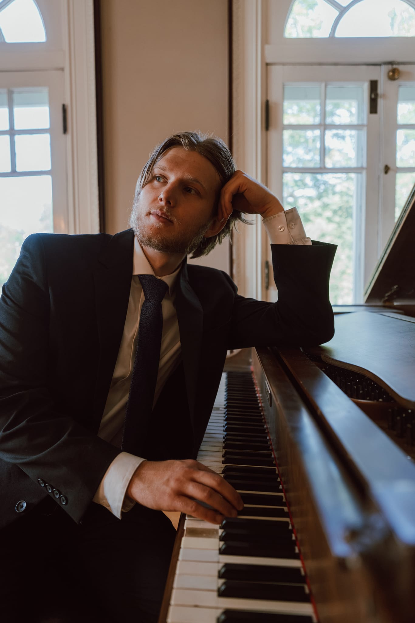 Abe in a suit resting his head on his hand at an upright piano, gazing upward