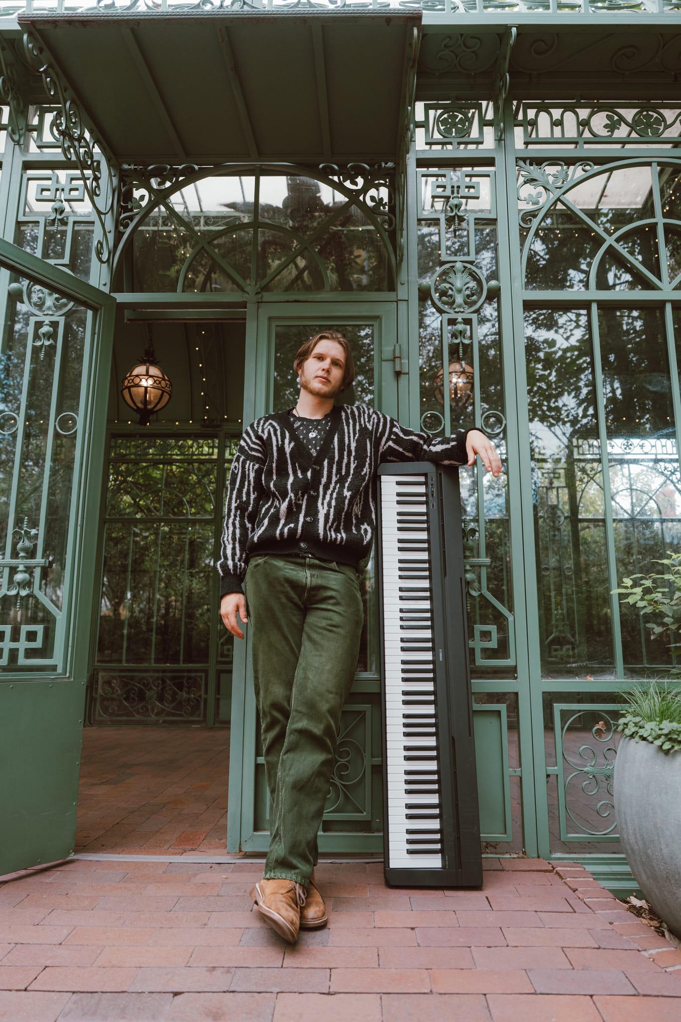 Abe standing in the doorway of an ornate green iron greenhouse, a digital keyboard propped beside him