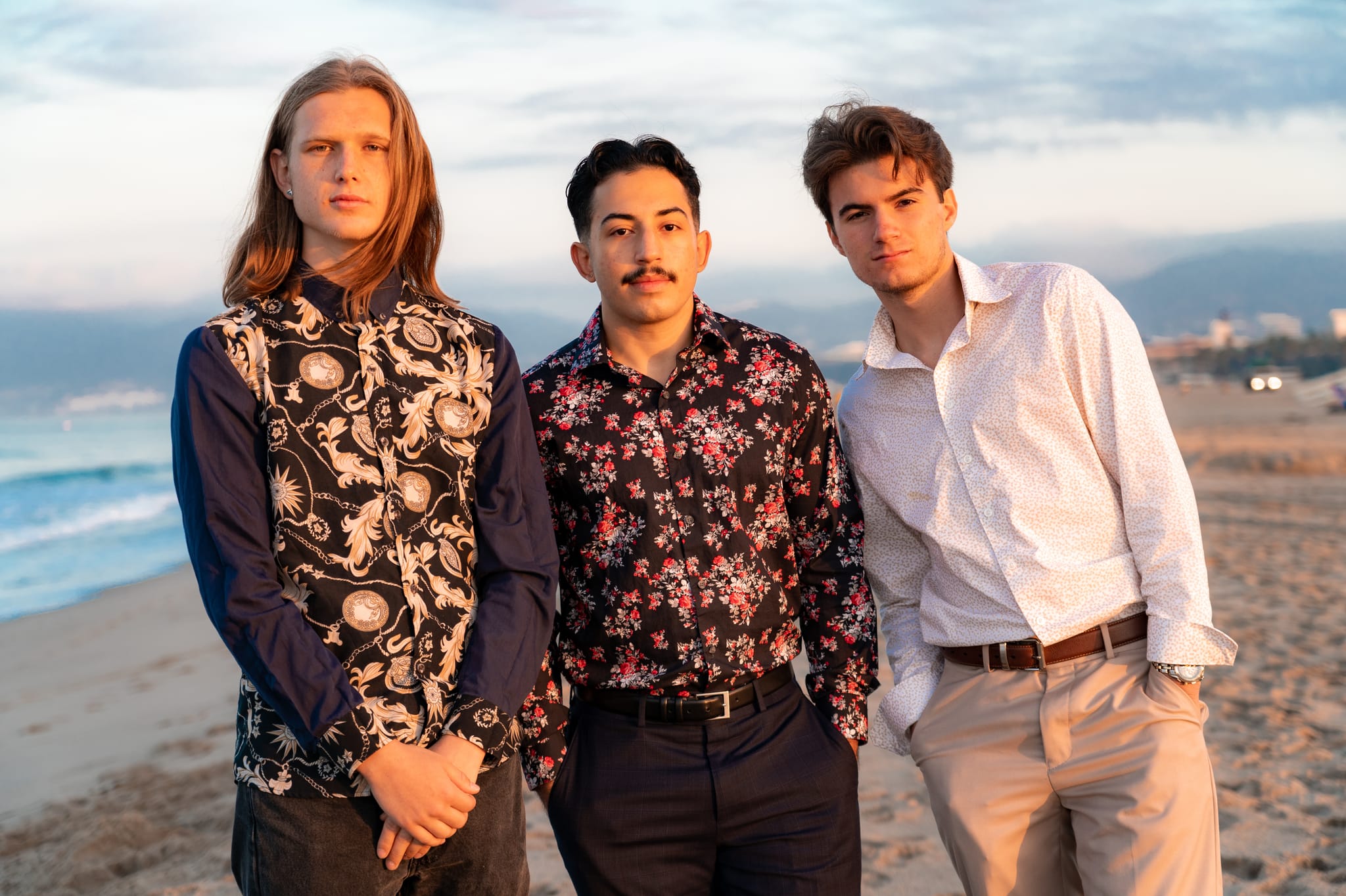 Abe and two bandmates in patterned shirts standing together on a beach at dusk